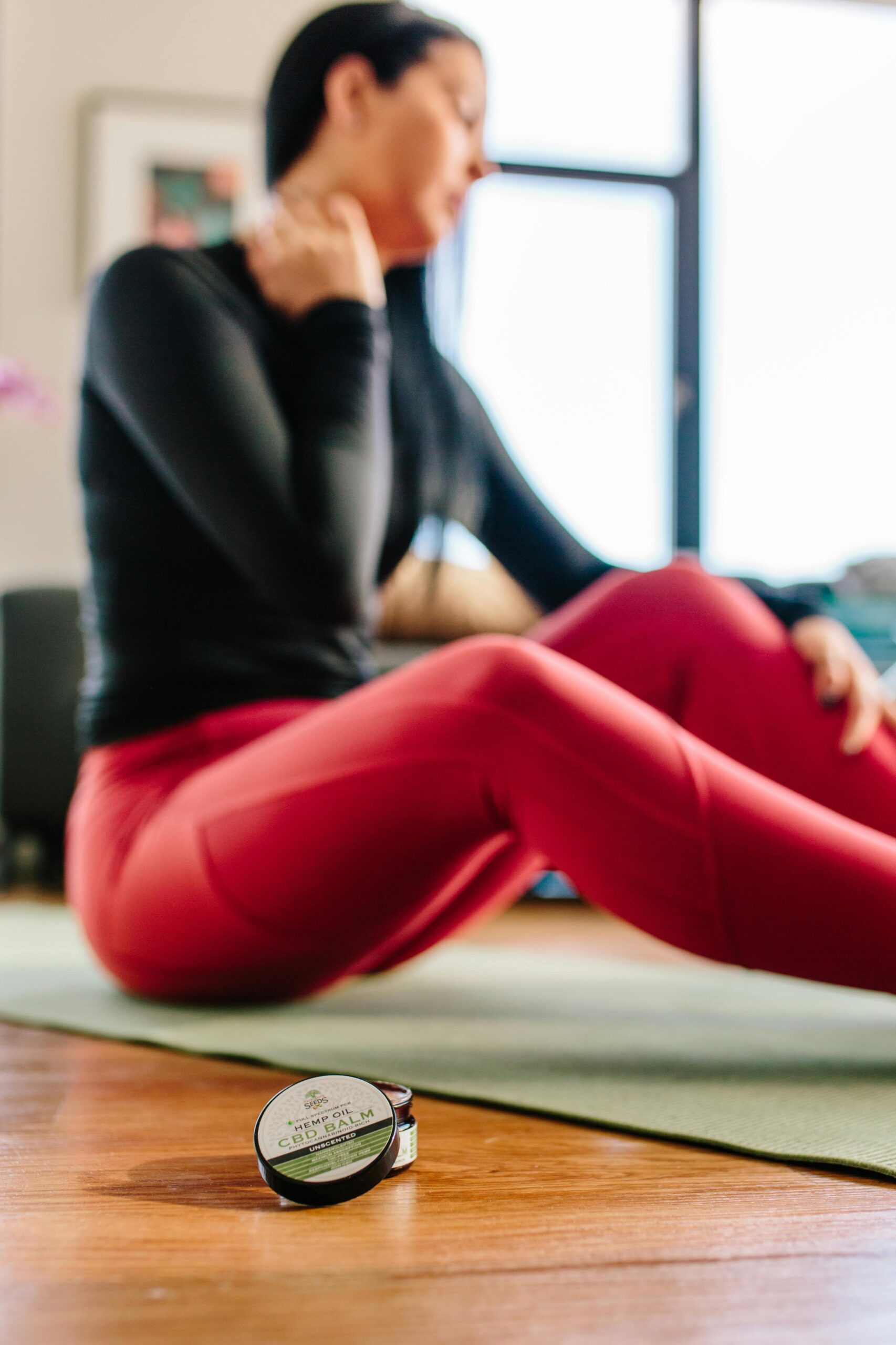 A woman sitting on a yoga mat indoors, using CBD cream for neck pain relief after exercise.