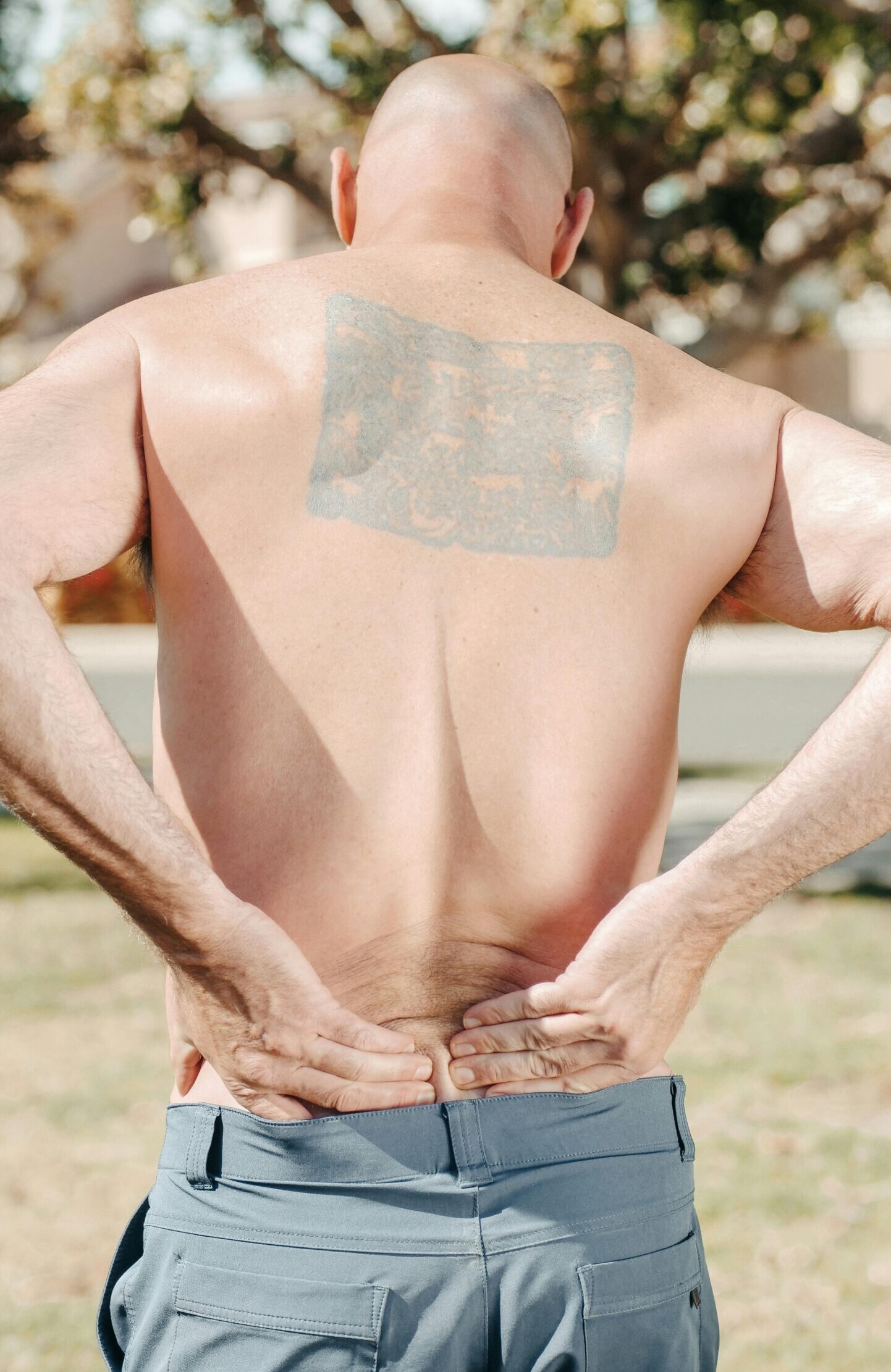 Shirtless bald man from behind holding his lower back, outdoors in sunny weather, with a large tattoo.